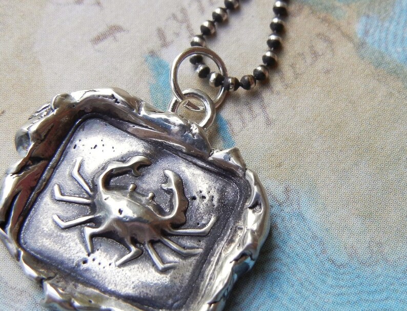 May include: A silver pendant necklace featuring a detailed crab design. The pendant is set in a square frame with a textured, aged finish. The chain is made of silver beads.