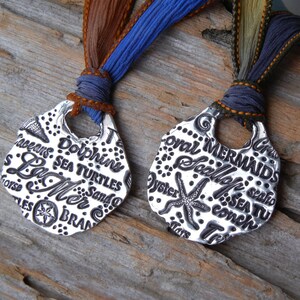 May include: Two silver pendants with a nautical theme. One pendant features the words "La Mer" and "Dolphins" and the other pendant features the words "Coral" and "Mermaids".