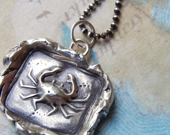 May include: A silver pendant necklace featuring a detailed crab design. The pendant is set in a square frame with a textured, aged finish. The chain is made of silver beads.