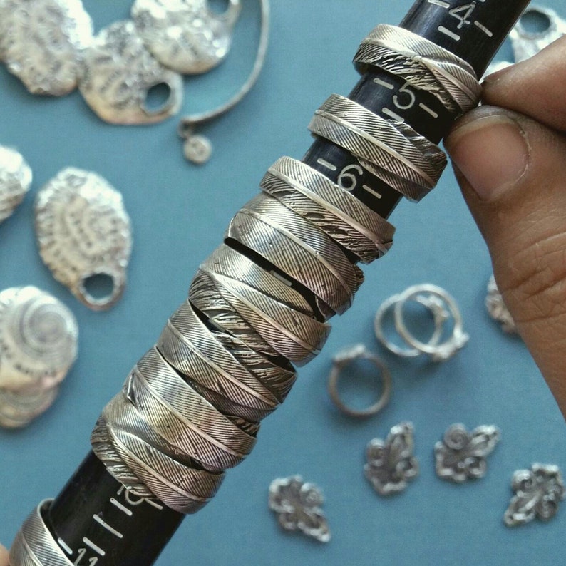 May include: A stack of silver rings with a textured feather design. The rings are stacked on a black ruler with white markings.
