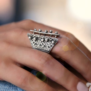 May include: A silver ring with a geometric design and a textured surface. The ring has a wide band and a raised, openwork section in the center.