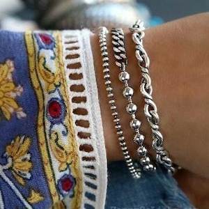 May include: Four silver bracelets stacked on a wrist. The top bracelet is a thick, twisted rope chain. The second bracelet is a thin, beaded chain. The third bracelet is a thin, plain chain. The bottom bracelet is a thick, beaded chain.