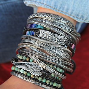 May include: A stack of silver bracelets with intricate designs, including a bracelet with the word "Dream" engraved on it, and a bracelet with a silver wing charm. The bracelets are layered on a person's wrist.
