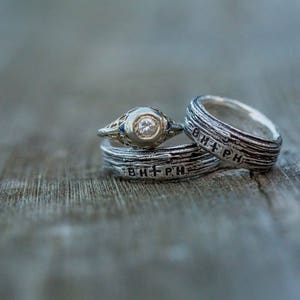 May include: Two silver wedding bands with a diamond engagement ring. The bands have the inscription "B+PH" on them.