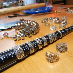 May include: A close-up of a silver ring sizing tool with a black ruler. The tool is made of metal rings of different sizes. The ruler is marked with numbers. There are several silver rings on the table.