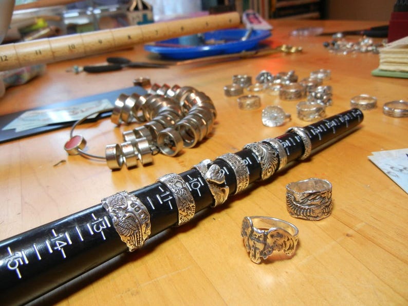 May include: A close-up of a silver ring sizing tool with a black ruler. The tool is made of metal rings of different sizes. The ruler is marked with numbers. There are several silver rings on the table.
