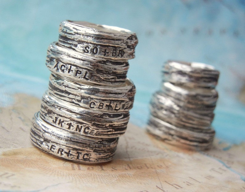 May include: A stack of silver rings with engraved initials and words, such as "SO+BR", "ACHPL", "CB+ER", "JK1+NC", and "ERT+TC".