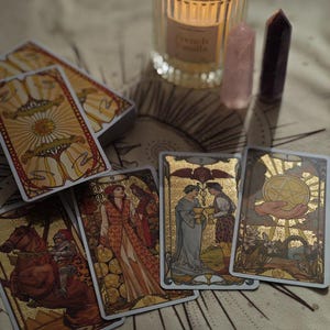 May include: A tarot card deck with gold accents and illustrations is displayed with several cards fanned out. A lit candle in a glass jar and two crystals are also present. The candle has a label that reads "French Vanilla".