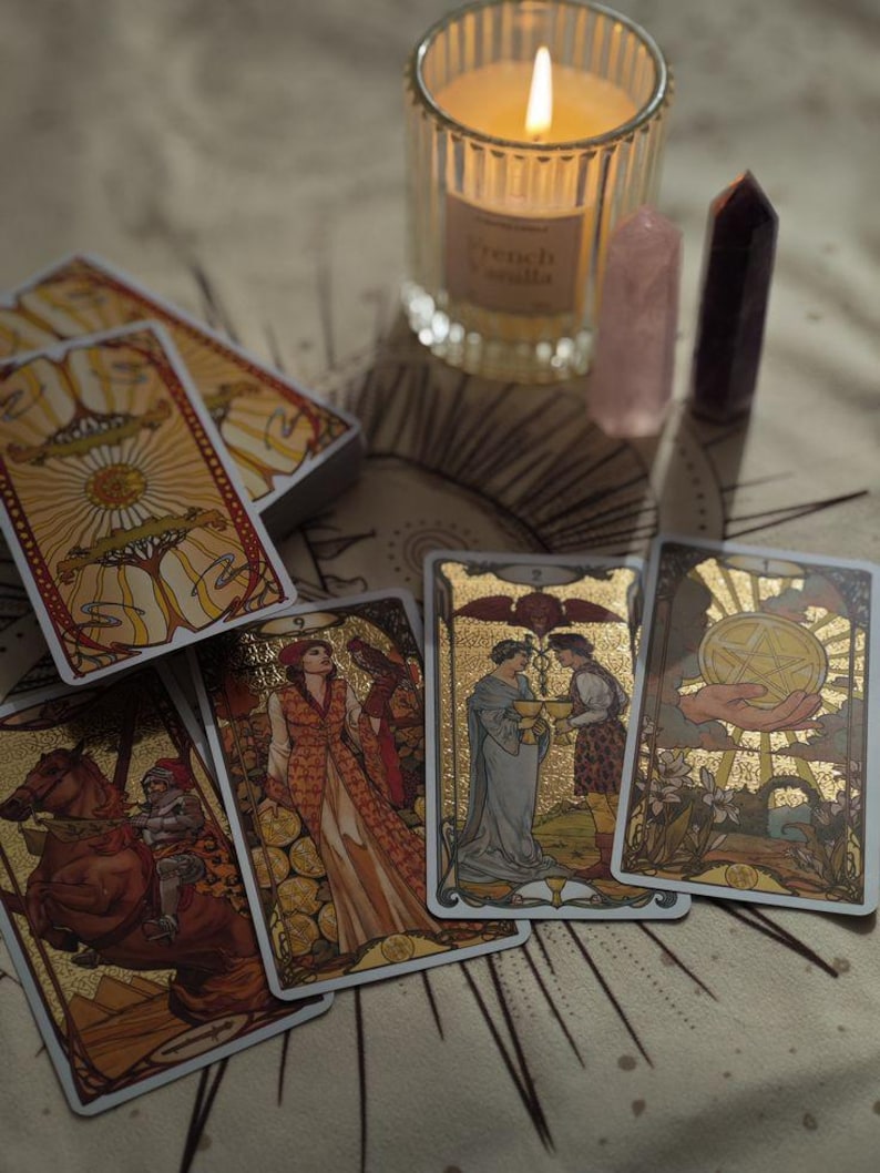 May include: A tarot card deck with gold accents and illustrations is displayed with several cards fanned out. A lit candle in a glass jar and two crystals are also present. The candle has a label that reads "French Vanilla".