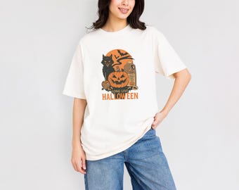 May include: Cream-colored t-shirt with a Halloween-themed graphic. The design features a black cat, a carved pumpkin, bats, and the text "LONG LIVE HALLOWEEN" in orange. The shirt is a casual, short-sleeved style.