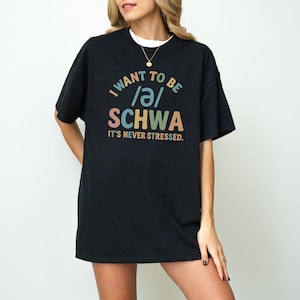 May include: Black t-shirt with the text "I want to be /ə/ SCHWA It's never stressed." The text is in a variety of colors, including blue, orange, and green. The shirt has short sleeves and a crew neck.