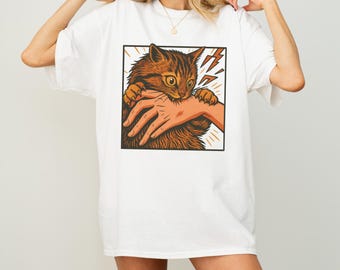 May include: White t-shirt featuring a cartoon graphic of a brown tabby cat biting a hand. The cat is inside a black square with yellow lightning bolts. The shirt is a casual, oversized fit.
