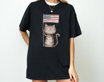 May include: Black t-shirt featuring a watercolor illustration of a cat holding an American flag. The cat is light brown with stripes and a smiling face. The flag has red and white stripes and a blue field with white stars. The shirt has a round neckline.