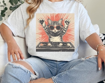 May include: A light grey t-shirt with a graphic of a cat wearing sunglasses and headphones, with its paws raised. The cat is behind a DJ mixing board, set against a red burst background.