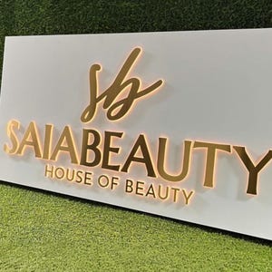 Könnte beinhalten: Ein weißes rechteckiges Schild mit goldfarbenem Schriftzug „SAIA BEAUTY HOUSE OF BEAUTY“. Die Buchstaben sind mit einem warmen Schein beleuchtet. Das Schild hat ein goldenes „sb“-Logo in der oberen linken Ecke.