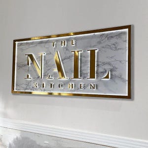 Puede incluir: Letrero dorado con fondo de mármol blanco. El letrero dice "The Nail Kitchen".