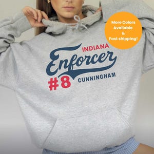Op de afbeelding: Een lichtgrijze hoodie met een voorzak en een capuchon met trekkoord. De hoodie toont de tekst "Indiana Enforcer #8 Cunningham" in marineblauw en rood. De woorden "Sport Grey" staan onderaan gedrukt.