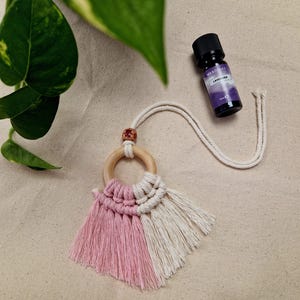 Könnte beinhalten: Ein rosa-weißer Makramee-Auto-Anhänger mit einem Holzring und einem Quasten. Der Anhänger hängt an einer weißen Schnur. Eine kleine Flasche Lavendel-Ätherisches Öl ist ebenfalls im Bild zu sehen.