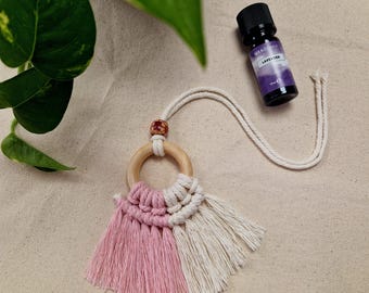 May include: A pink and white macrame car charm with a wooden ring and a tassel. The charm is hanging from a white cord. A small bottle of lavender essential oil is also in the image.