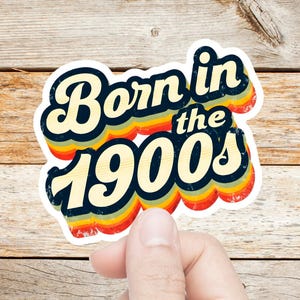 May include: A die-cut sticker featuring the text Born in the 1900s in a bold, dark blue font. The lettering is layered over a series of horizontal, wavy stripes in shades of yellow, orange, and red, creating a multi-colored, retro-inspired design.