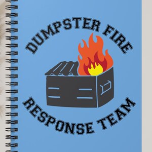 May include: A spiral-bound notebook with a light blue cover. The cover features a black dumpster graphic with flames and the text "DUMPSTER FIRE RESPONSE TEAM" in a black, arched font. The notebook is open.