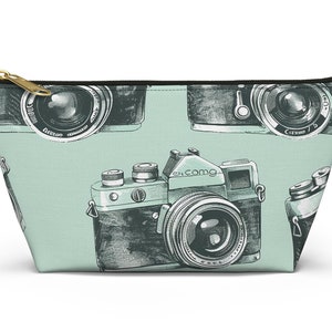 May include: A zippered pouch with a light mint green background featuring a repeating pattern of black and white vintage camera illustrations. The pouch has a gold-toned metal zipper pull at the top and a black fabric trim along the upper edge.