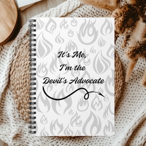 May include: A white spiral bound notebook with a gray flame pattern and the text "It's Me, I'm the Devil's Advocate" in black.