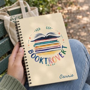 May include: A spiral-bound notebook with a cream-colored cover featuring a watercolor illustration of a stack of books topped with an open book. The text "BOOKTROVER" is arched in colorful letters, and the name "Carrie" is printed in blue script.