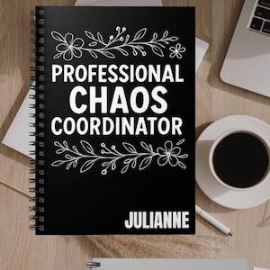 May include: Black spiral-bound notebook with the text "PROFESSIONAL CHAOS COORDINATOR" in white, adorned with floral and leaf designs. The name "JULIANNE" is printed at the bottom. A cup of coffee and a laptop are visible in the background.