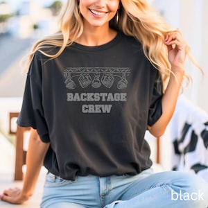 May include: Black t-shirt with the text "BACKSTAGE CREW" in a white, stylized font. Above the text is a graphic of stage lights. The shirt is a dark color and has a relaxed fit.