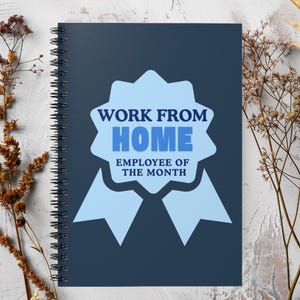 Könnte beinhalten: Ein marineblaues Spiralnotizbuch mit dem Text "WORK FROM HOME EMPLOYEE OF THE MONTH" in einem hellblauen Abzeichen-Design. Das Notizbuch ist von getrockneten Pflanzen umgeben.