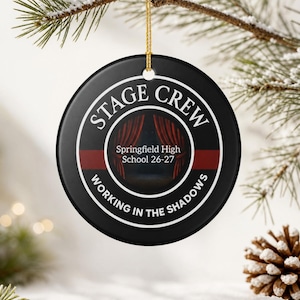 May include: A round black ceramic ornament featuring a white circular border with the text STAGE CREW and WORKING IN THE SHADOWS. The center displays red stage curtains and the text Springfield High School 26-27, suspended by a gold string.