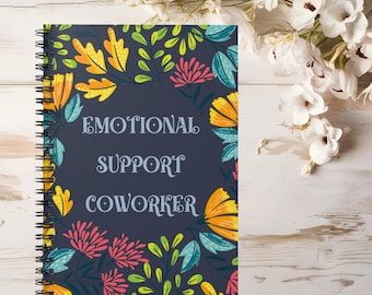 May include: A spiral-bound notebook with a dark navy blue cover featuring a colorful floral pattern of yellow, pink, and teal leaves and flowers. The text "EMOTIONAL SUPPORT COWORKER" is printed in a light gray serif font in the center.