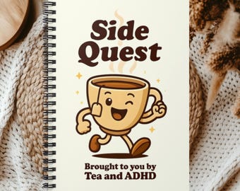 May include: A spiral-bound notebook with a cream-colored cover featuring a cartoon tea cup character with arms and legs. The text reads Side Quest at the top and Brought to you by Tea and ADHD at the bottom in dark brown, bold, rounded lettering.