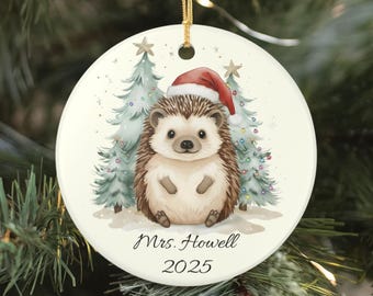 May include: A round white ceramic Christmas ornament featuring a brown hedgehog wearing a red Santa hat, positioned between two light green pine trees decorated with colorful lights. The text Mrs. Howell and the year 2025 are printed below.