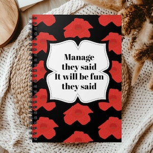 May include: A spiral-bound notebook with a black cover featuring a repeating pattern of red flowers. In the center, a white scalloped-edge label contains the black text: Manage they said It will be fun they said. The notebook has a black wire coil.