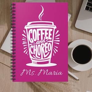 May include: A magenta spiral-bound notebook with a white coffee cup graphic and the words "COFFEE and CHOREO." The name "Ms. Maria" is below. A cup of coffee and a laptop are on the wooden surface beside the notebook.