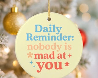 May include: A round, pale yellow ceramic ornament hanging from a gold string. The text reads Daily Reminder: nobody is mad at you in blue, peach, and red lettering, accented with small star and flower symbols printed on the front surface.