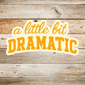 May include: A sticker with the phrase "a little bit DRAMATIC" in orange lettering. The text is outlined in a cream color and set against a wooden background.