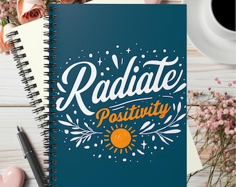 Może przedstawiać: Ciemnoniebieski notatnik spiralny z białym napisem "Radiate" nad pomarańczowym napisem "Positivity", któremu towarzyszy grafika stylizowanego pomarańczowego słońca, białych gałązek z liśćmi oraz małych białych gwiazdek na okładce.