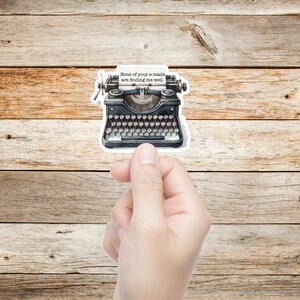 Op de afbeelding: Een sticker van een zwarte typemachine in vintage-stijl met een wit vel papier waarop de tekst "None of your e-mails are finding me well." is gedrukt. De typemachine heeft een gedetailleerd toetsenbord, ronde toetsen en metalen accenten.
