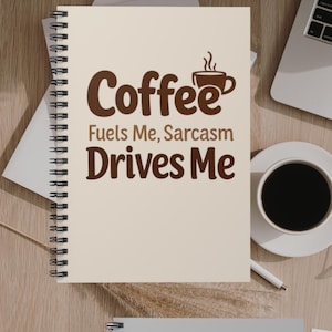 May include: A spiral-bound notebook with the words "Coffee Fuels Me, Sarcasm Drives Me" in brown lettering. A coffee cup graphic is above the word "Coffee". A cup of coffee and a laptop are in the background.