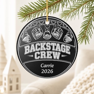 May include: A round black ceramic ornament featuring a white graphic of stage lights and the text BACKSTAGE CREW. Below the graphic, the name Carrie and the year 2026 are printed in white. The ornament hangs from a gold string.