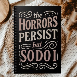 Pode incluir: Caderno preto com espiral com o texto "the HORRORS PERSIST but SO DO I" em letras rosa e brancas. O caderno está aberto, mostrando páginas em branco, e repousa sobre uma manta de malha creme.