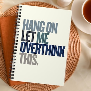 Peut inclure: Un carnet à spirales blanc avec le texte "HANG ON LET ME OVERTHINK THIS." en lettres bleues, grises et blanches.