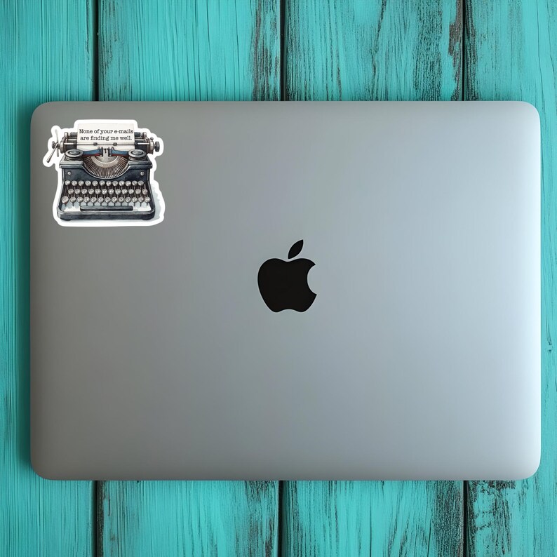 Op de afbeelding: Een zilveren laptop met een zwart Apple-logo in het midden en een sticker van een zwarte vintage typemachine in de linkerbovenhoek. De typemachinesticker bevat een wit vel papier met de tekst None of your e-mails are finding me well.