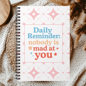 May include: A spiral-bound notebook with a white cover featuring a repeating pink diamond and dot pattern. The center displays the text Daily Reminder: nobody is mad at you in blue, orange, and red, accented by small star and flower icons.