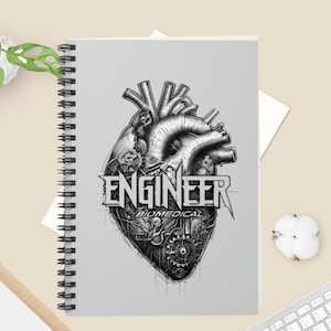 May include: A spiral-bound notebook with a light gray cover featuring a black and white illustration of a mechanical human heart integrated with gears and wires. The text ENGINEER BIOMEDICAL is printed in a bold, stylized font across the center.