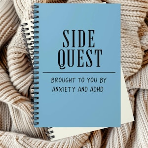 以下が含まれることがあります： 黒い文字で「SIDE QUEST」と書かれた青いスパイラルノート。テキストの下には、黒い文字で「BROUGHT TO YOU BY ANXIETY AND ADHD」と書かれています。