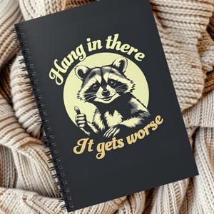 May include: A black spiral notebook with a raccoon illustration and the text "Hang in there It gets worse".